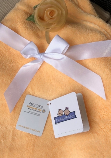 Flauschige orange Babydecke mit weißer Satinschleife und zarter, hellgelber Stoffrose. Zwei Anhänger liegen auf der Decke: Oeko-Tex Standard 100 Zertifikat und KiddiBaby-Label. Sie zeigen Sicherheit und Komfort.