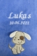 Azurblaue, superflauschige Babydecke, bestickt mit dem Namen Lukas und dem Geburtsdatum 10.06.2021 in weißer Schrift. Darunter ein applizierter beiger Hund mit weißen Schlappohren und braunem Fleck. Freundlich, kindgerecht.